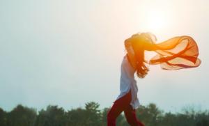 Foto de mulher de perfil com blusa branca, calça vermelha, cabelos compridos e segurando um véu esvoaçante contra o pôr-do-sol