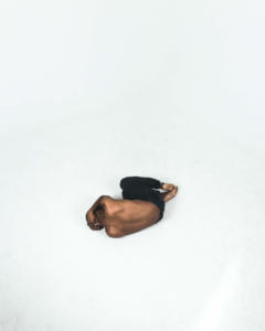 Foto afastada de um homem negro sem camisa deitado em posição fetal sobre um chão totalmente branco