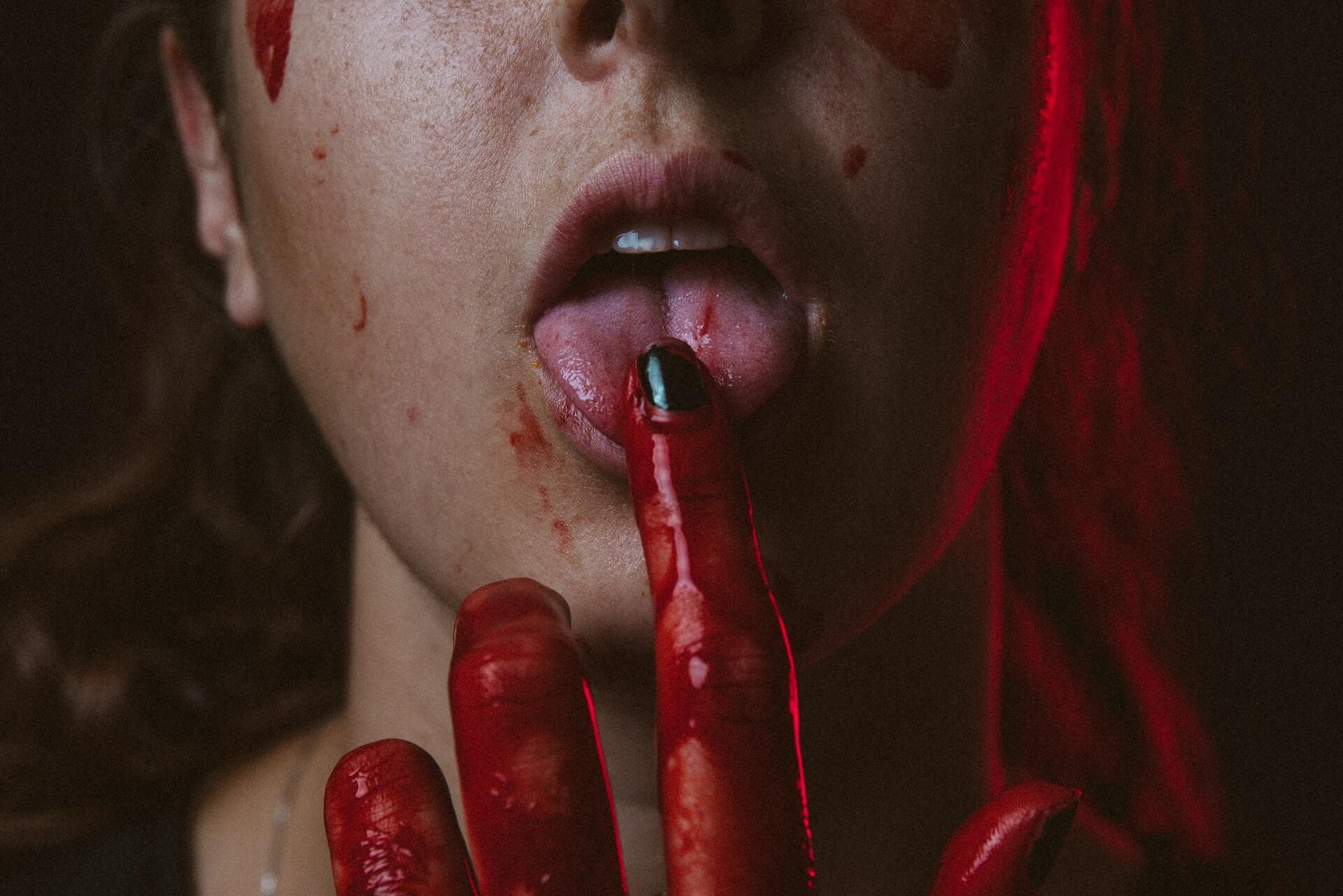 Foto aproximada da boca de uma mulher com o indicador na ponta da língua, com a unha pintada de preto e a mão toda ensanguentada