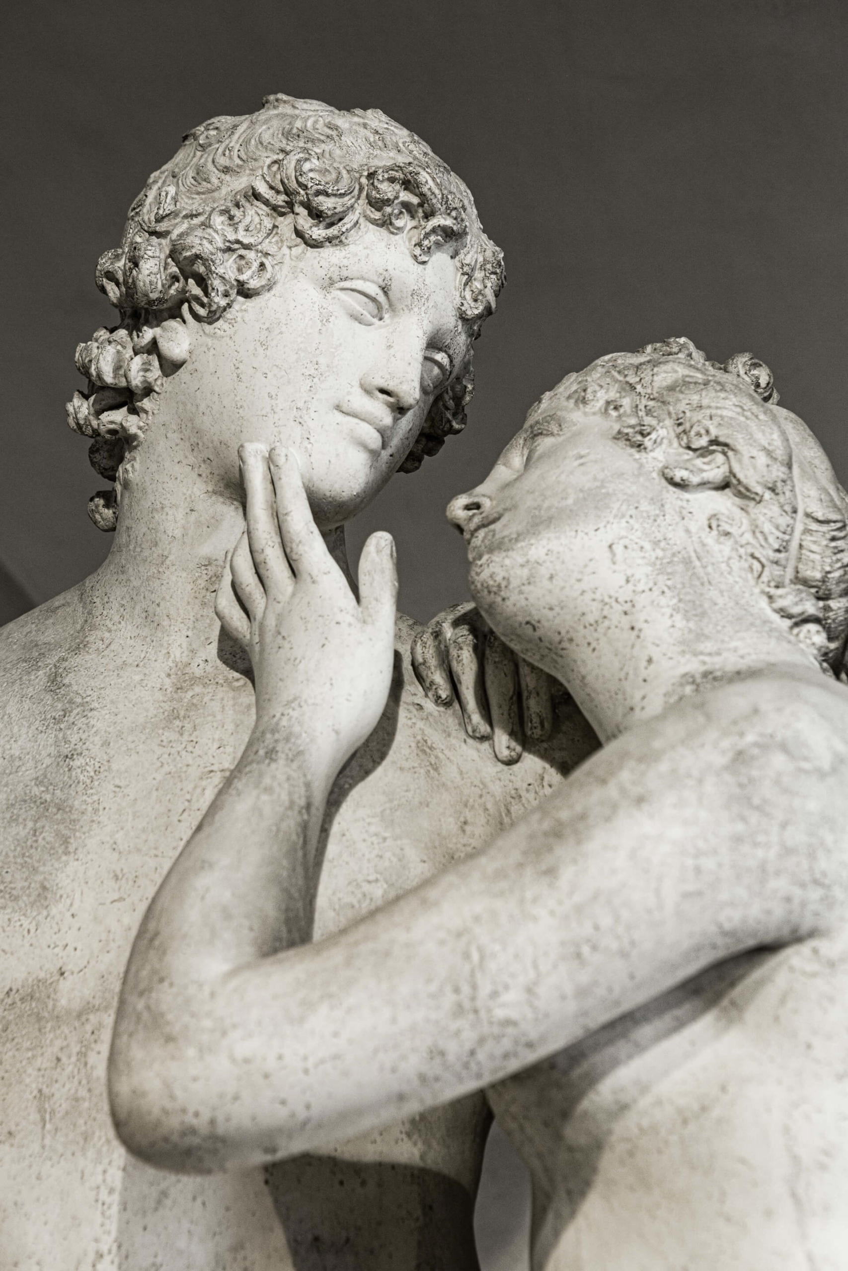 Foto detalhada da estátua de um casal: uma pessoa se apoiando delicadamente sobre o ombro da outra, com a mão tocando levemente sua face, e as duas se olhando apaixonadamente
