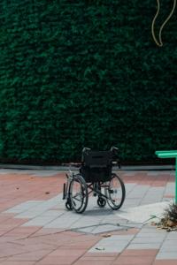 Foto de uma cadeira de rodas vazia com uma bengala sobre o assento, estacionada em um pátio com chão de placas de cimento alternadas entre cinza e avermelhado, em frente a uma cerca viva de plantas bem viçosas
