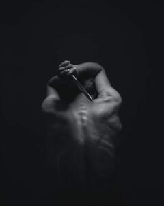 Foto em preto e branco de um homem de costas, sem camisa, segurando uma adaga prateada com as duas mãos sobre a cabeça