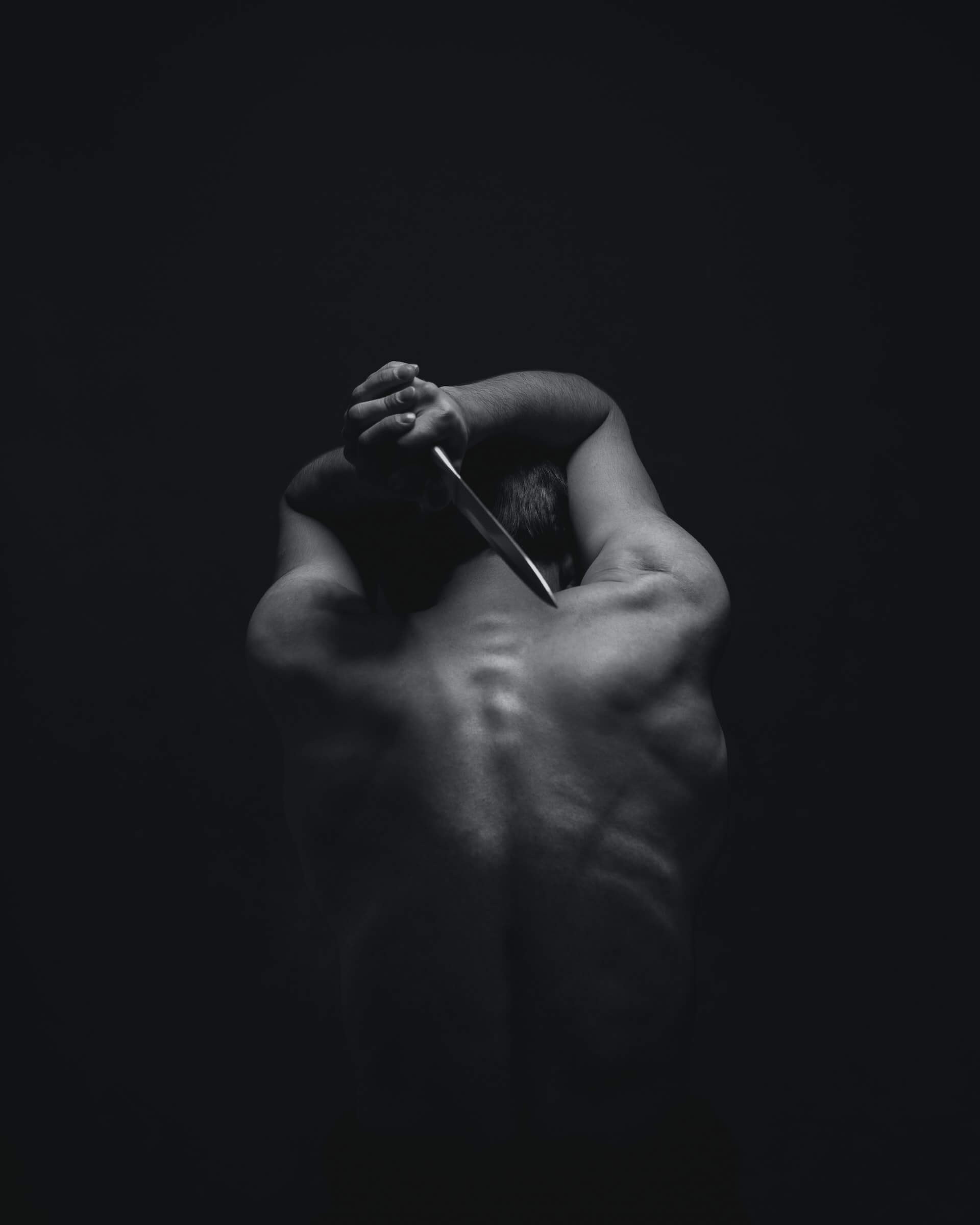 Foto em preto e branco de um homem de costas, sem camisa, segurando uma adaga prateada com as duas mãos sobre a cabeça