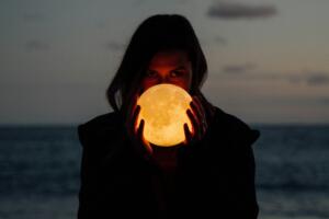Foto de uma mulher de cabelos lisos compridos com o mar desfocado ao fundo e segurando em frente ao rosto, na altura da boca e do nariz, uma luminária acesa esférica com textura de solo lunar