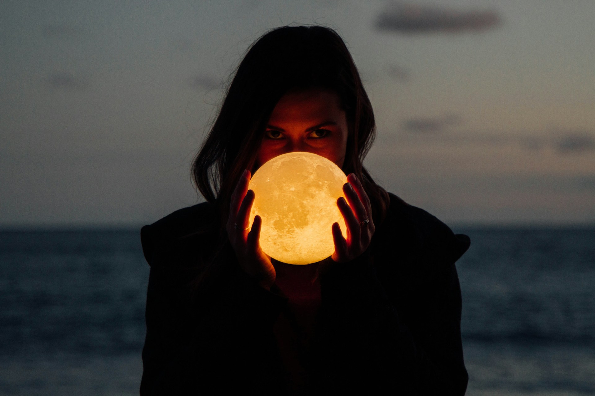 Foto de uma mulher de cabelos lisos compridos com o mar desfocado ao fundo e segurando em frente ao rosto, na altura da boca e do nariz, uma luminária acesa esférica com textura de solo lunar