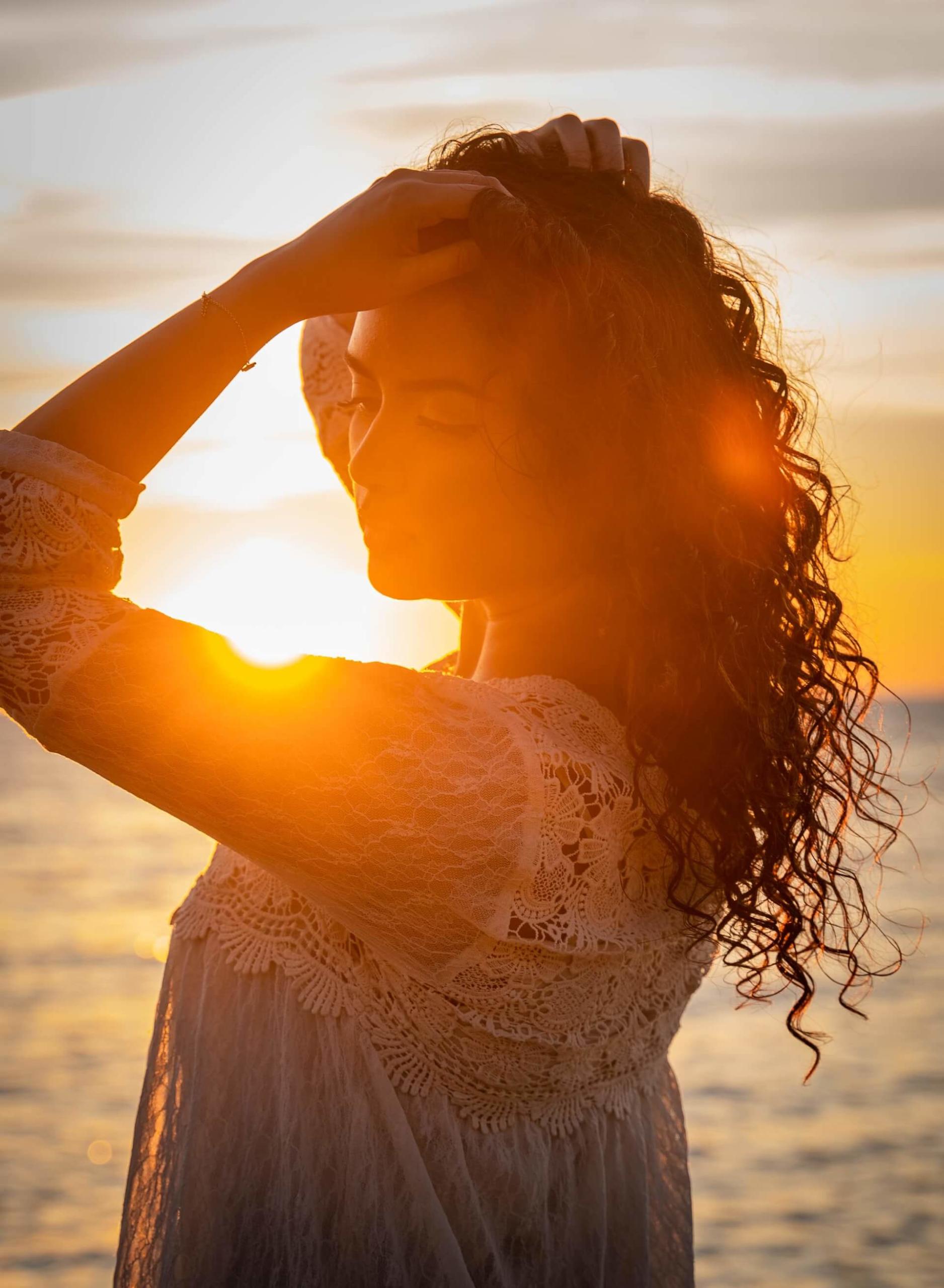Foto da silhueta de uma mulher com um vestido de renda branca massageando os próprios cabelos levemente ondulados contra o pôr-do-sol
