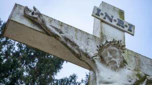 Foto aproximada de um detalhe de uma estátua da crucificação de Jesus em concreto branco, mostrando o rosto com a coroa de espinhos, a mão direita pregada à cruz, e a placa colocada acima da cabeça com a inscrição "I.N.R.I."