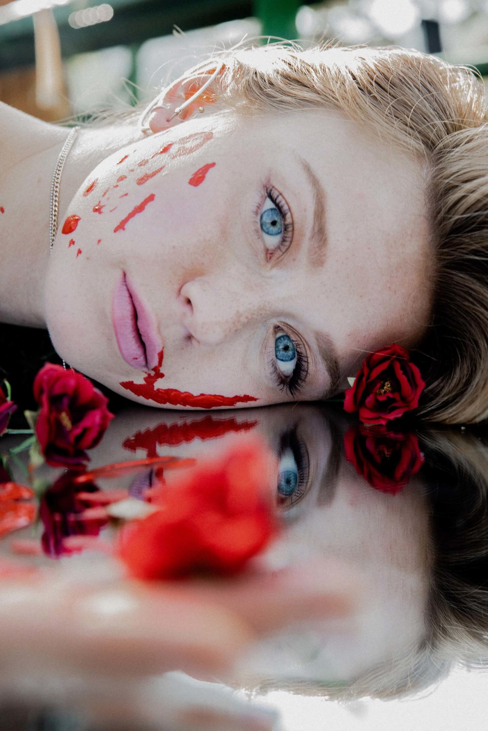 Foto aproximada e macabra do rosto ensanguentado de uma mulher loira com olhos azuis bem claros, deitada entre algumas rosas vermelhas sobre uma superfície bem reflexiva