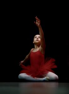 Foto de uma bailarina com vestido vermelho, meia calça branca, sapatilha clara, sentada no chão com as pernas cruzadas e o braço esquerdo estendido para cima