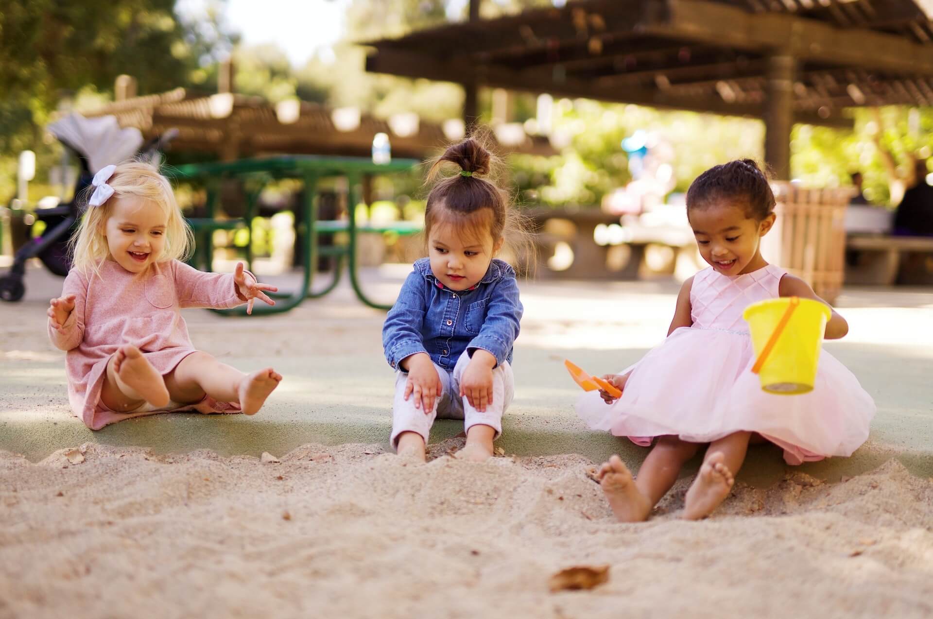 Foto de três meninas bem pequenas, sorrindo e brincando com os pés num banco de areia