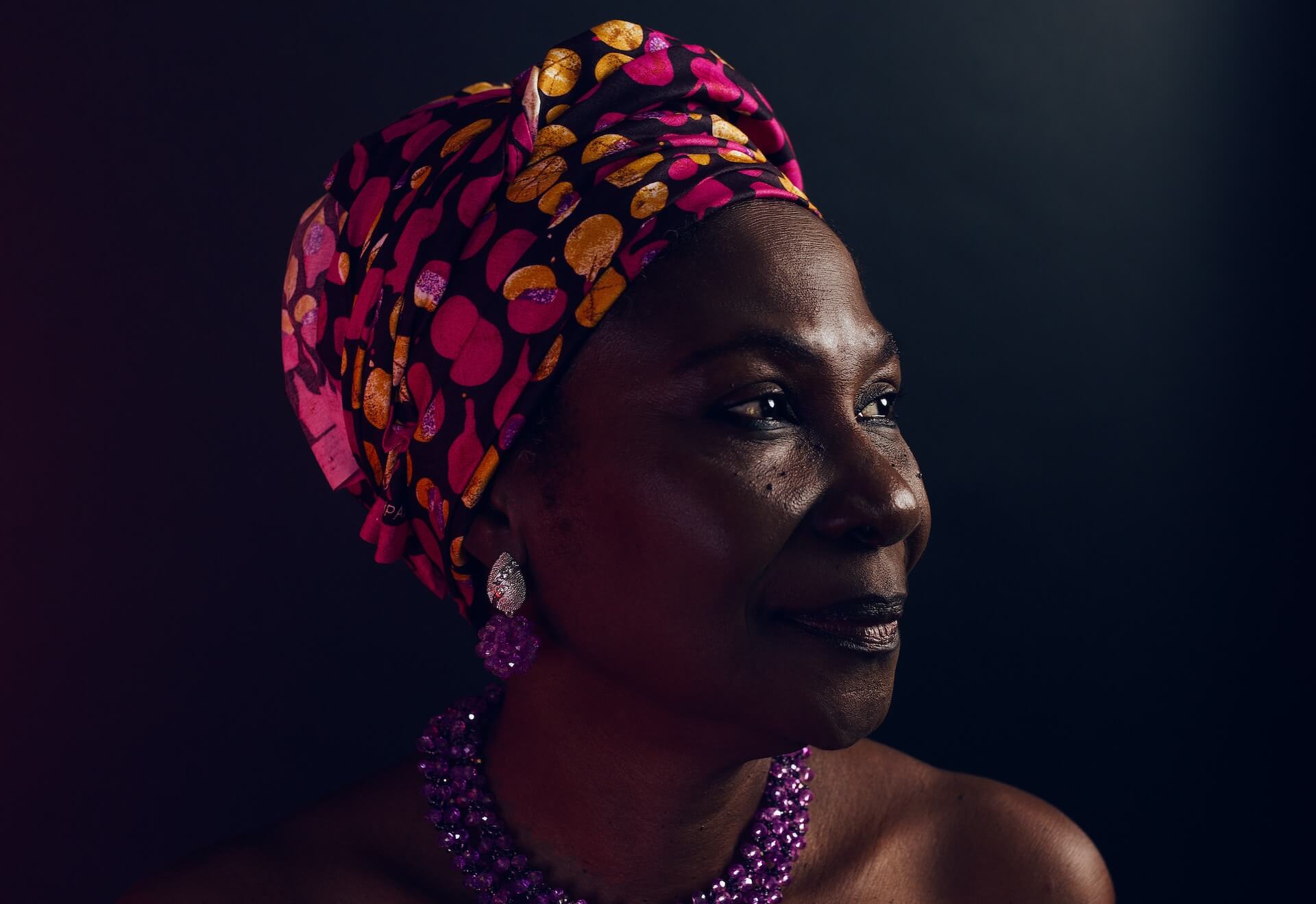 Retrato de perfil de uma mulher preta com turbante colorido e brinco e colar roxos cintilantes