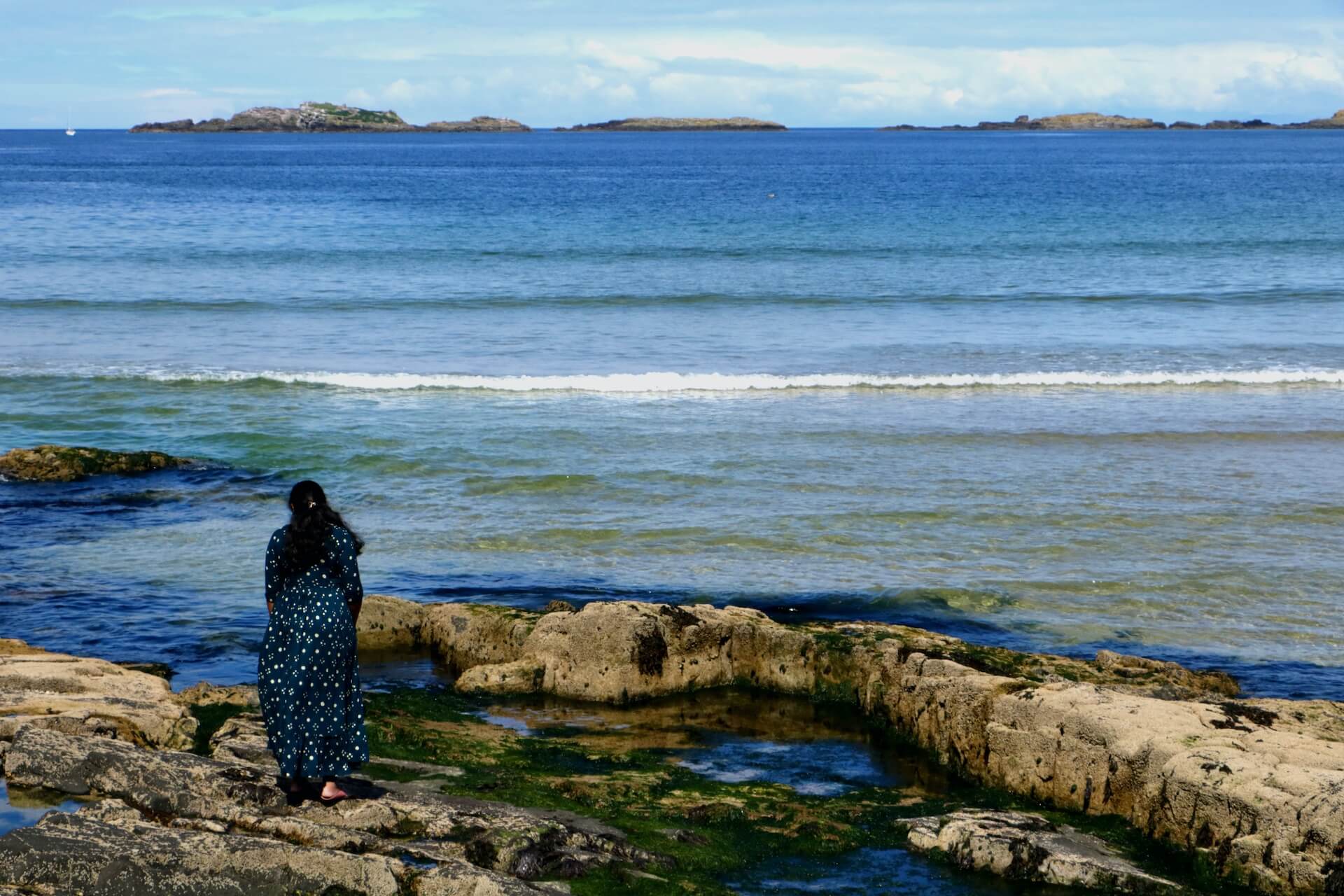 Foto de uma mulher de costas, usando um vestido estampado azul, de pé em uma costa rochosa na beira do oceano