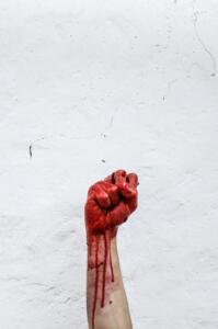 Foto de um punho erguido banhado em sangue em frente a uma parede rústica branca com algumas rachaduras