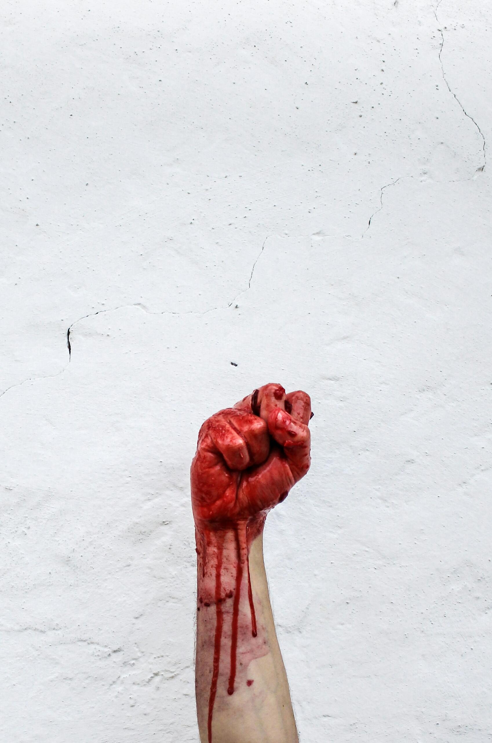 Foto de um punho erguido banhado em sangue em frente a uma parede rústica branca com algumas rachaduras