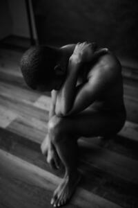 Foto em preto e branco de um homem negro nu ajoelhado, de cabeça baixa, com uma das mãos no pescoço, sobre um piso de madeira.