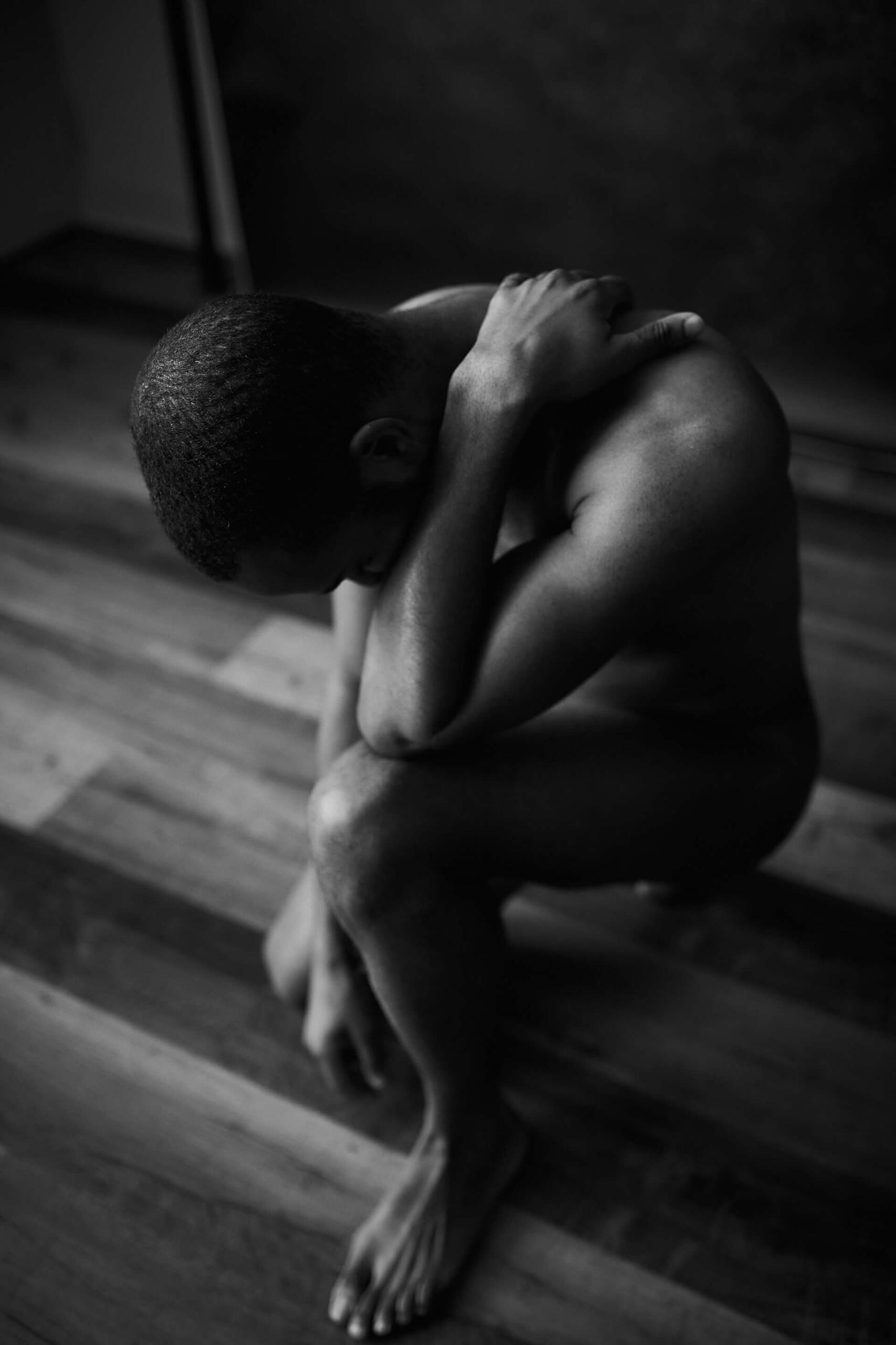 Foto em preto e branco de um homem negro nu ajoelhado, de cabeça baixa, com uma das mãos no pescoço, sobre um piso de madeira.